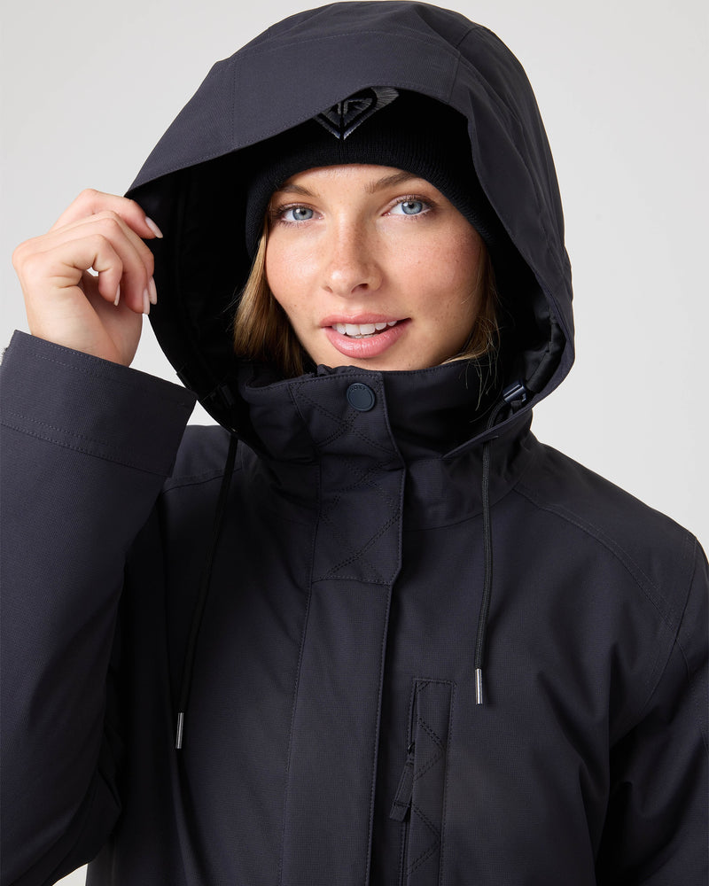 A person wearing the Roxy Billie Jacket in black and a black beanie, smiling and holding the edge of the hood with one hand, against a plain background.