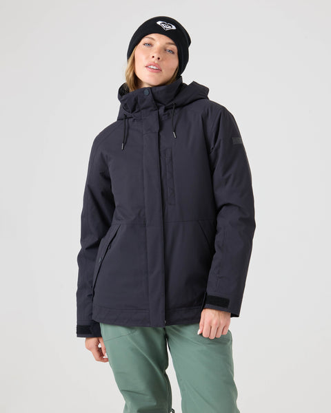 A person wearing the Roxy Billie Jacket in black, paired with green pants and a black beanie featuring a white logo, stands against a plain light background.