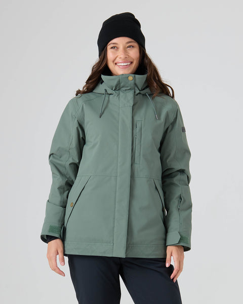 A smiling person wears the Roxy Billie Jacket in Duck Green, paired with black pants and a black beanie, standing against a plain light background.