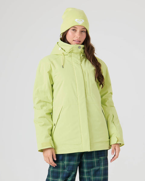 A woman wearing the Roxy Billie Jacket in Lily Green, paired with a matching beanie and blue-green plaid pants, stands against a plain background, looking at the camera with a neutral expression.