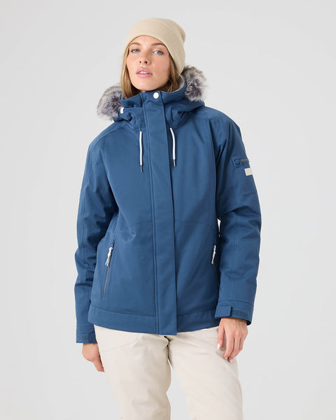A person wearing a beige beanie, light pants, and a Roxy Meade Jacket in Ensign Blue with a faux fur-lined hood stands against a plain gray background.