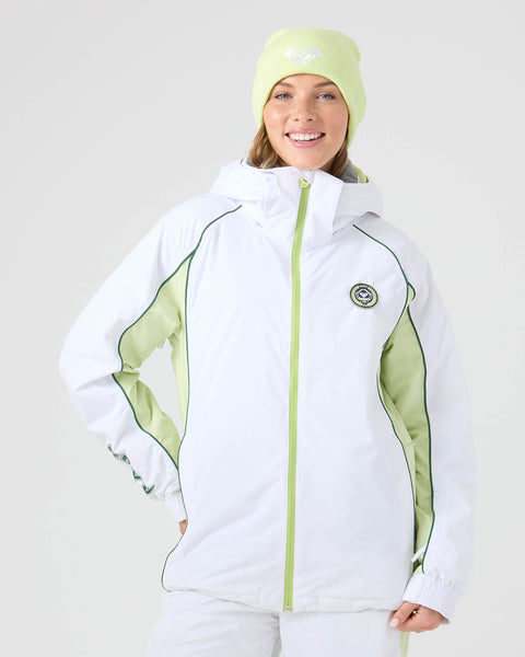 A smiling woman stands against a plain background, wearing a light green beanie and the Roxy Chloe Kim Honors Jacket in Bright White, featuring green trim and a circular chest patch. This women’s snowboard jacket is made from recycled polyester, combining style with sustainability.