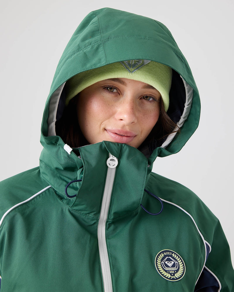 A person wearing the Roxy Chloe Kim Honors Jacket, a green hooded women’s snowboard jacket with mapped warmth and a crest, paired with a light green beanie, looks at the camera with a neutral expression against a plain background.