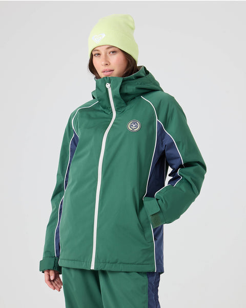 A person wearing the Roxy Chloe Kim Honors Jacket—a green women’s snowboard jacket featuring premium weatherproofing and mapped warmth—paired with green pants and a light yellow beanie, stands against a plain light background, looking at the camera.