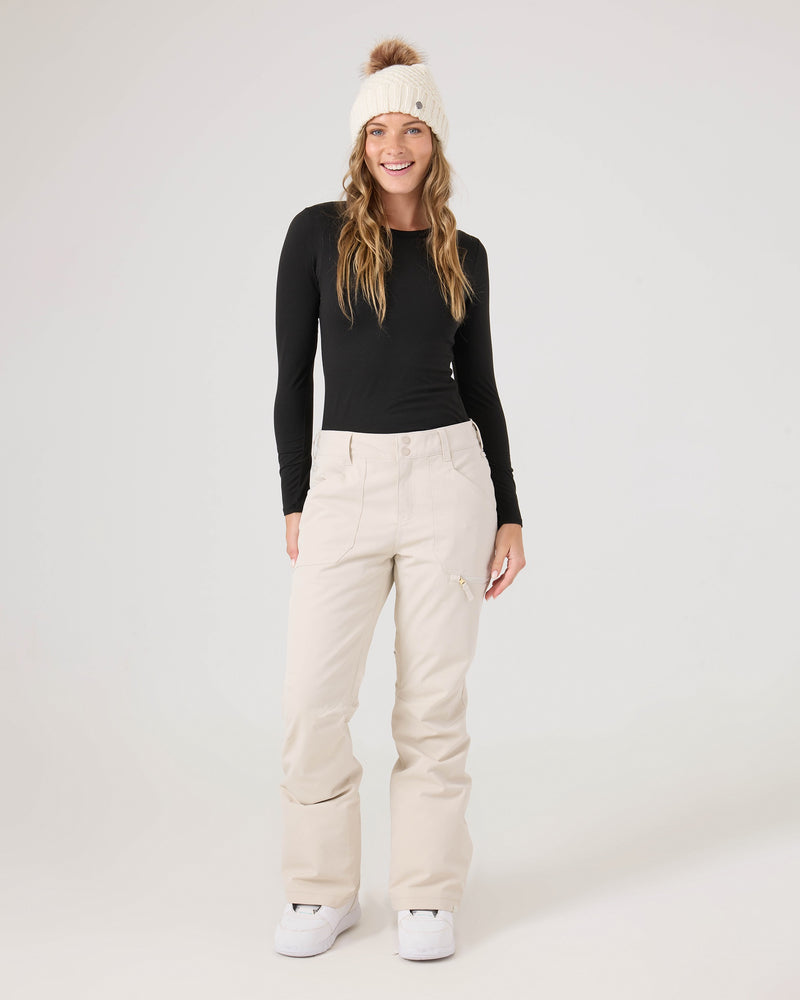A woman in a black long-sleeve top, Roxy’s Nadia Pants in beige with a tailored fit, and a white knit hat stands smiling against a plain light backdrop.