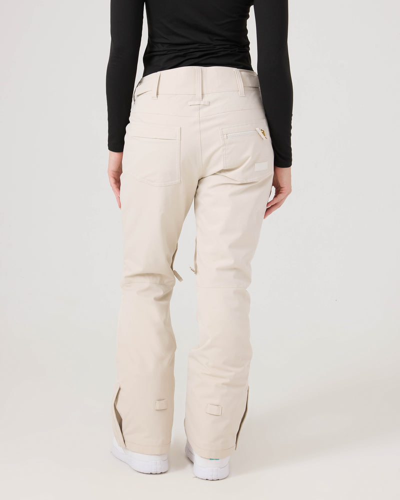 A person is seen from behind wearing a black long-sleeve top and Roxy Nadia Pants in cream, showcasing the tailored fit, back pockets, and flared legs of the women’s snow pants over white shoes.