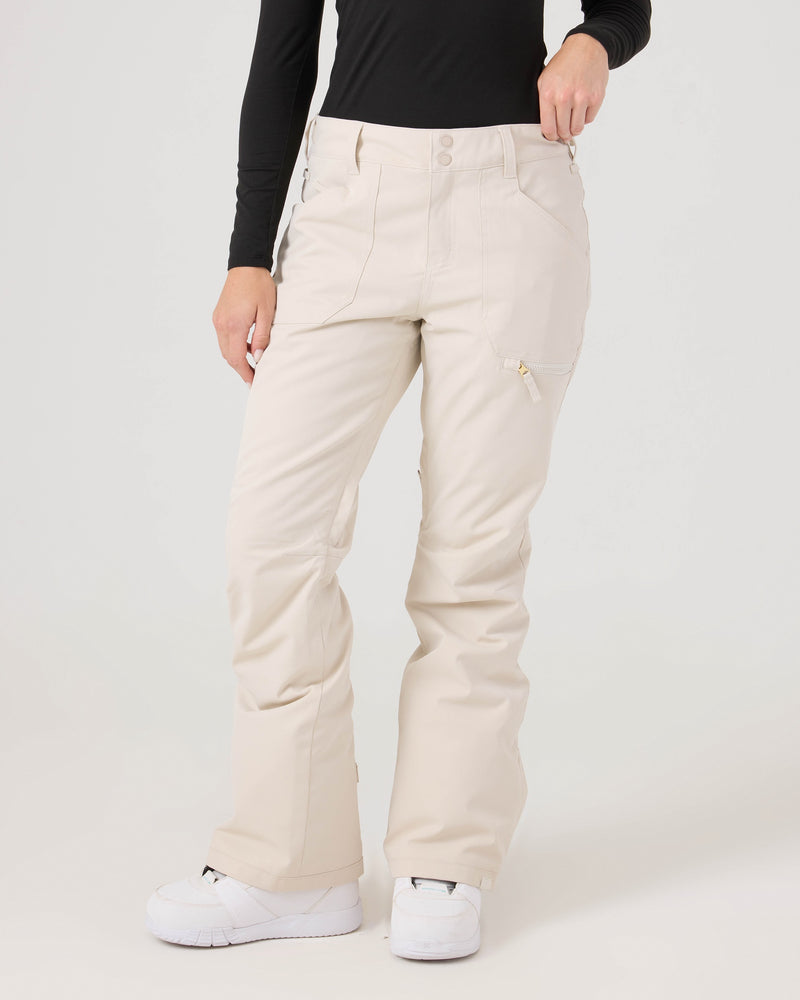 A person wearing Roxy Nadia Pants in Moonbeam—a pair of cream-colored women’s snow pants with a tailored fit—along with a black long-sleeve top and white shoes, stands against a plain light background. The Nadia Pants provide reliable 10K/10K protection for added comfort and style.