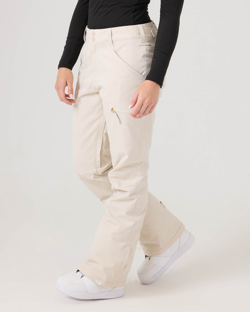 A person wearing a black long-sleeve shirt and the Roxy Nadia Pants in light beige, featuring a tailored fit and a zippered thigh pocket, paired with white athletic shoes, stands against a plain light gray background.