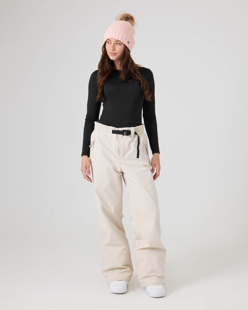 A woman stands against a plain light background wearing a pink knit beanie, a long-sleeve black top, and Roxy’s Bleeker Pants in loose beige with a belt—these women’s snow pants combine both style and comfort.
