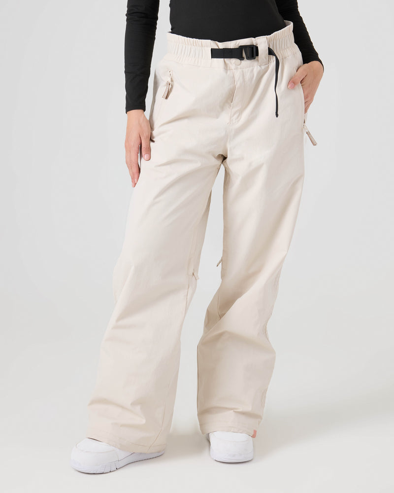 A person is wearing Roxy Bleeker Pants in a loose fit, light beige color, paired with a black belt, a black long-sleeve top, and white sneakers. Their left hand rests in their pocket. The plain light background emphasizes the 10K/5K protection offered by these women’s snow pants.