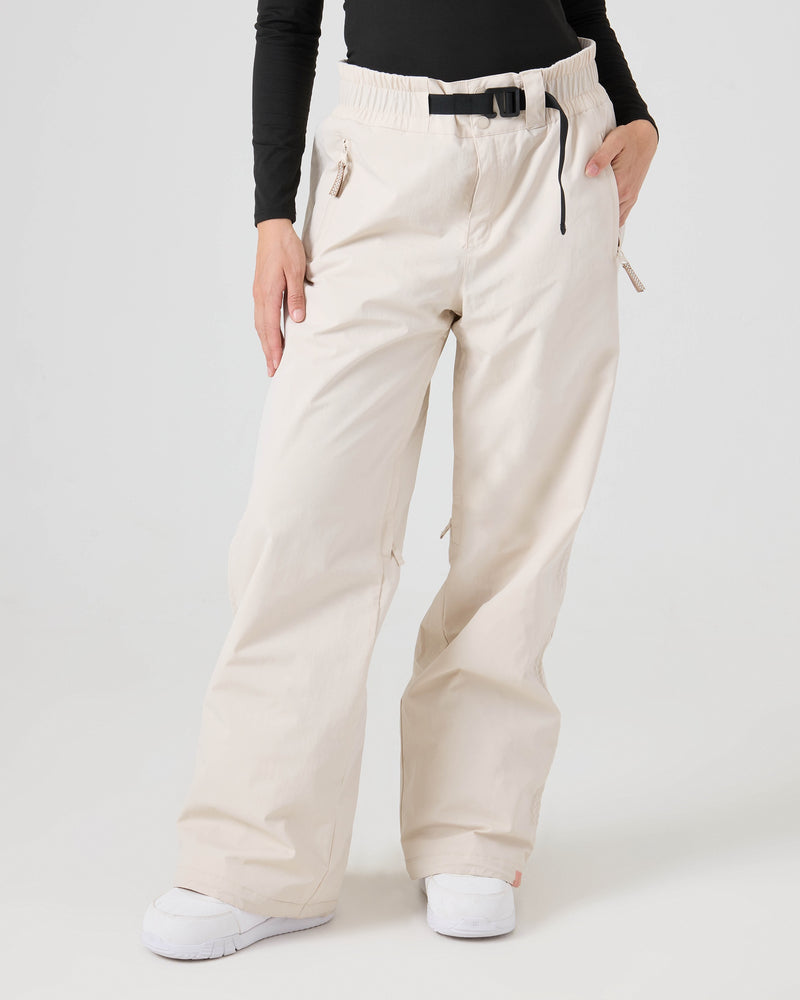 A person wearing wide-leg, light beige Roxy Bleeker Pants, which feature zippered pockets and a black belt, paired with a black long-sleeve top and white sneakers, stands against a plain white background. These women’s snow pants from Roxy provide 10K/5K protection for both style and performance.