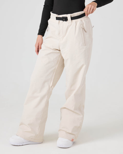 A person wearing Roxy Bleeker Pants in Moonbeam—off-white, loose-fitting women’s snow pants with 10K/5K protection—paired with a black belt, black long-sleeve top, and white shoes stands against a plain white background.