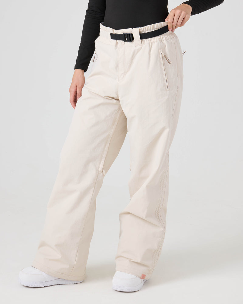 A person wearing Roxy Bleeker Pants in Moonbeam—off-white, loose-fitting women’s snow pants with 10K/5K protection—paired with a black belt, black long-sleeve top, and white shoes stands against a plain white background.