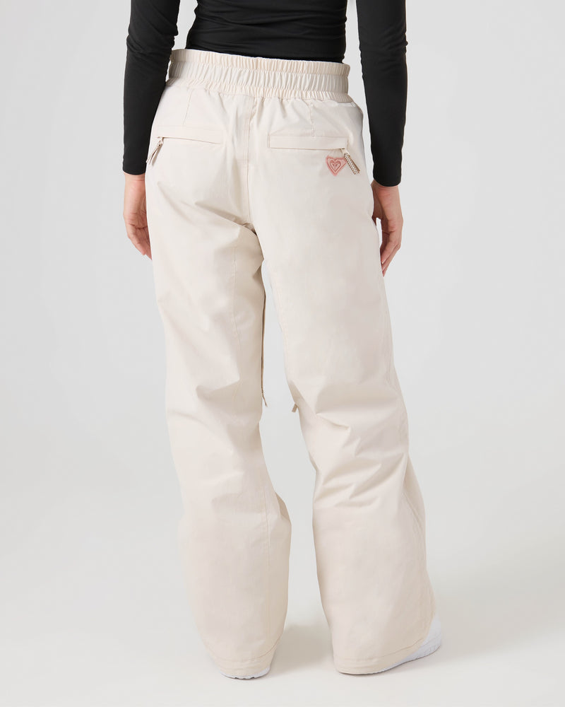 A person is shown from behind wearing loose-fitting, high-waisted Roxy Bleeker Pants in light beige, featuring back pockets and a small stitched pink heart detail. These women’s snow pants offer 10K/5K protection for comfort and style. The outfit is paired with a fitted black long-sleeve top.