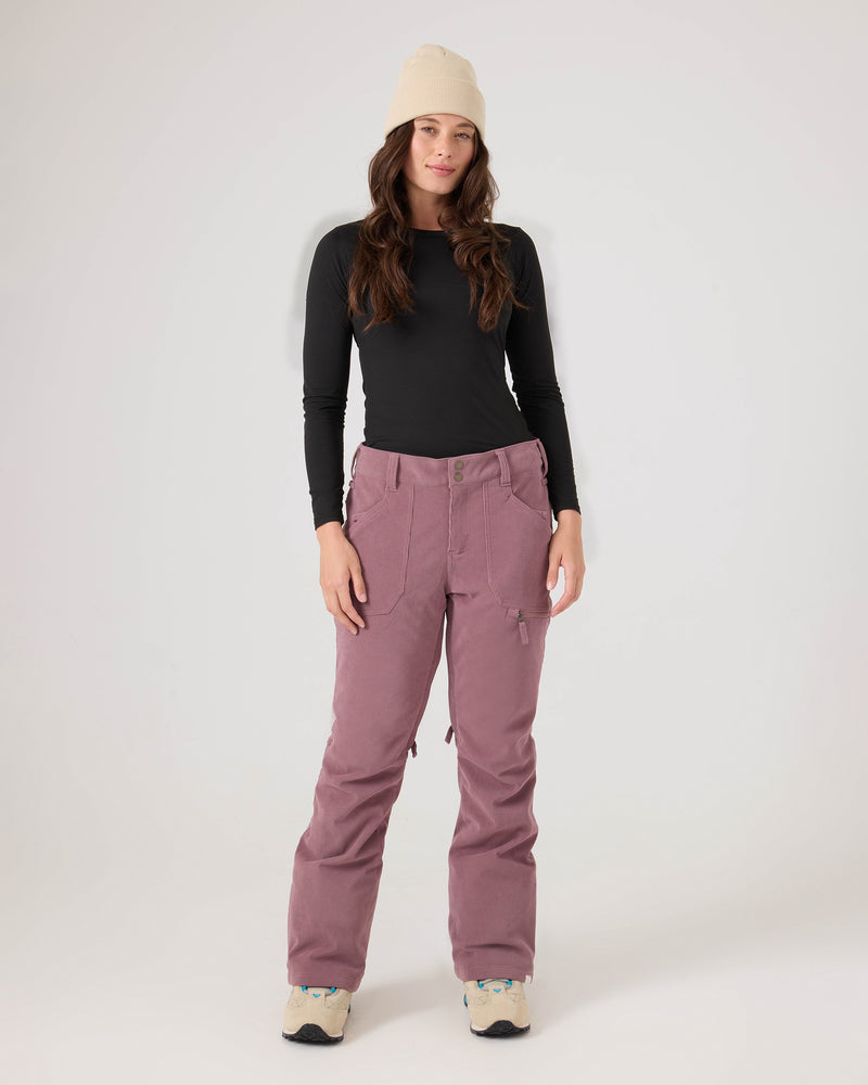 A person wearing a beige beanie, black long-sleeve shirt, Nadia Pants by Roxy in mauve, and sneakers stands against a plain white background, looking at the camera with a relaxed expression.