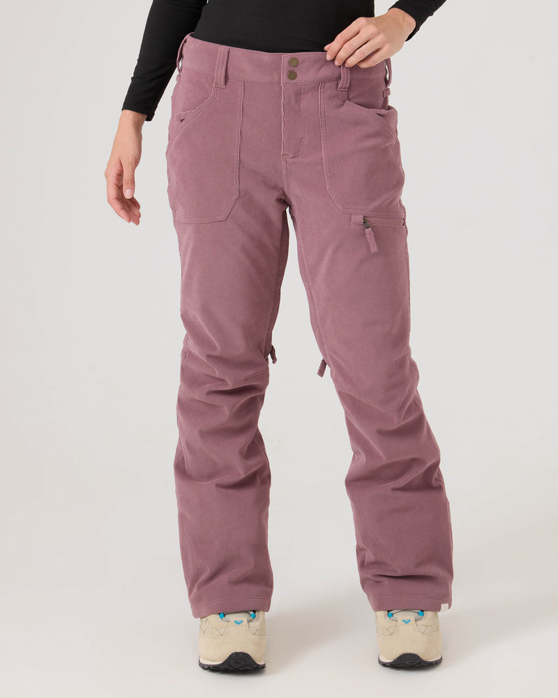 A person is shown wearing the Nadia Pants by Roxy in a mauve corduroy fabric with a tailored fit, paired with a black long-sleeve top. Only the lower body and one arm are visible as they stand against a plain light background. The outfit is completed with beige shoes featuring blue accents.