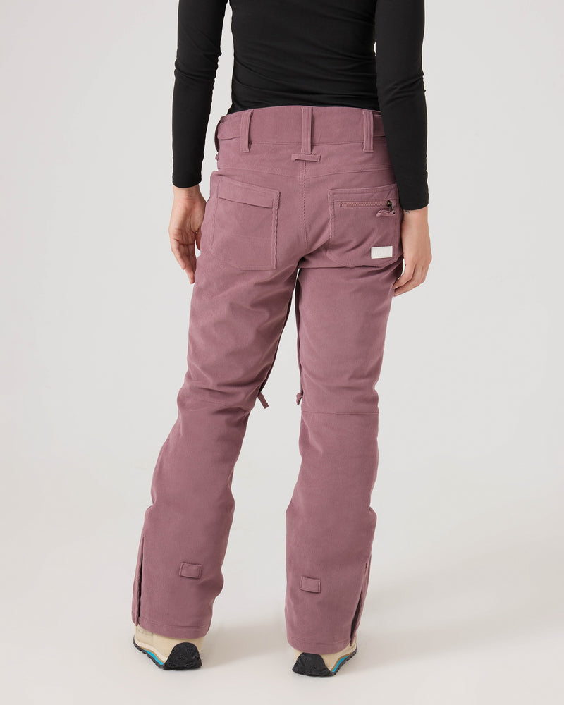 A person is seen from the back wearing the Nadia Pants by Roxy—mauve-colored, tailored-fit corduroy pants inspired by women’s snow pant styling. Featuring back pockets, adjustable hems, and crafted from recycled polyester, the outfit is paired with a black long-sleeve shirt. The scene is set against a plain, light background.