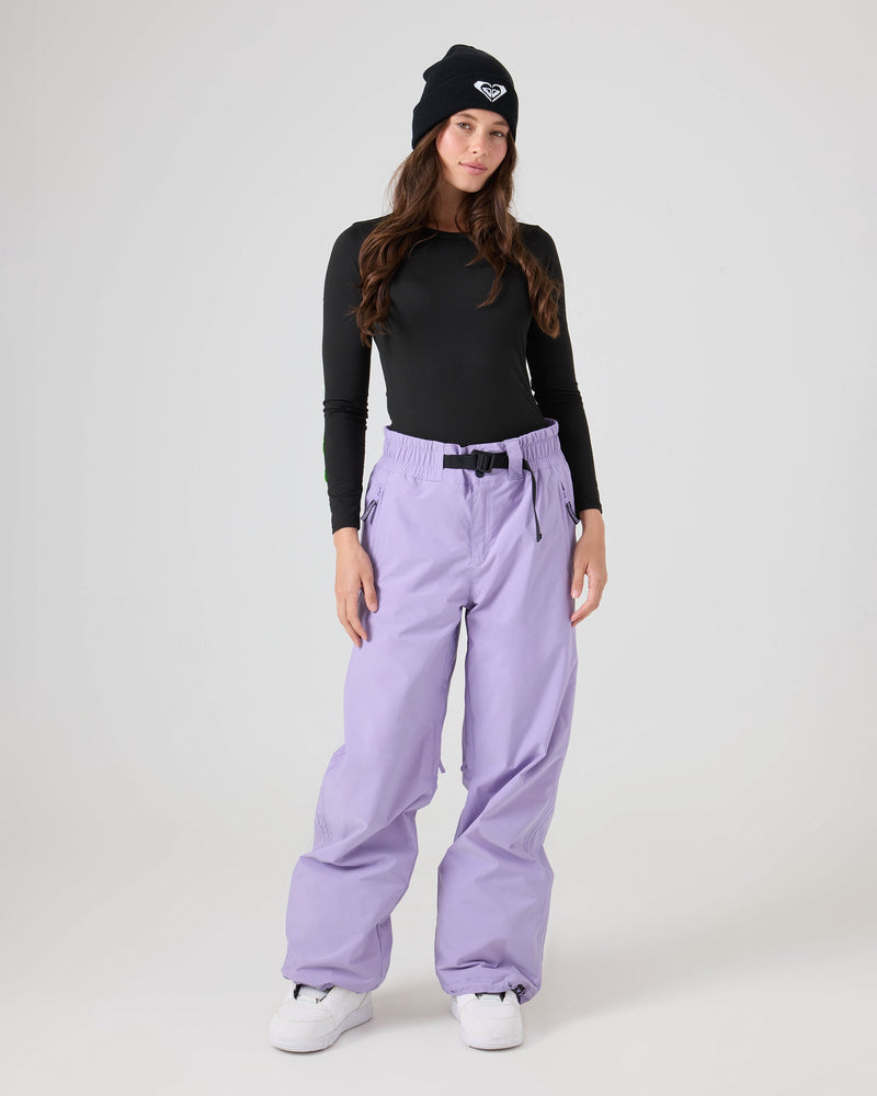 A person wearing a black long-sleeve shirt, Roxy Bleeker Pants in lilac with 10K/5K protection, white sneakers, and a black beanie stands against a plain light background.