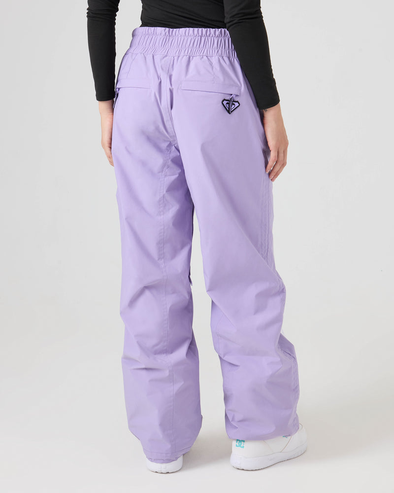 A person is seen from behind wearing Roxy Bleeker Pants in lavender, which offer 10K/5K weather protection and subtle venting for added comfort. The look is finished with a black long-sleeve top and white shoes, while an embroidered Roxy logo appears on the back pocket of the pants.