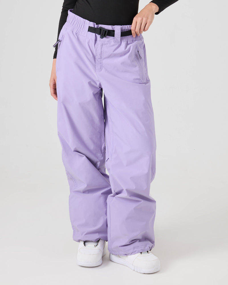 A person is shown from the waist down wearing Roxy Bleeker Pants—loose-fitting women’s snow pants in lavender with a black belt, zipper pockets, venting details, and an adjustable hem—paired with a black long-sleeve top and white sneakers. The pants offer 10K/5K weather protection for enhanced resistance.