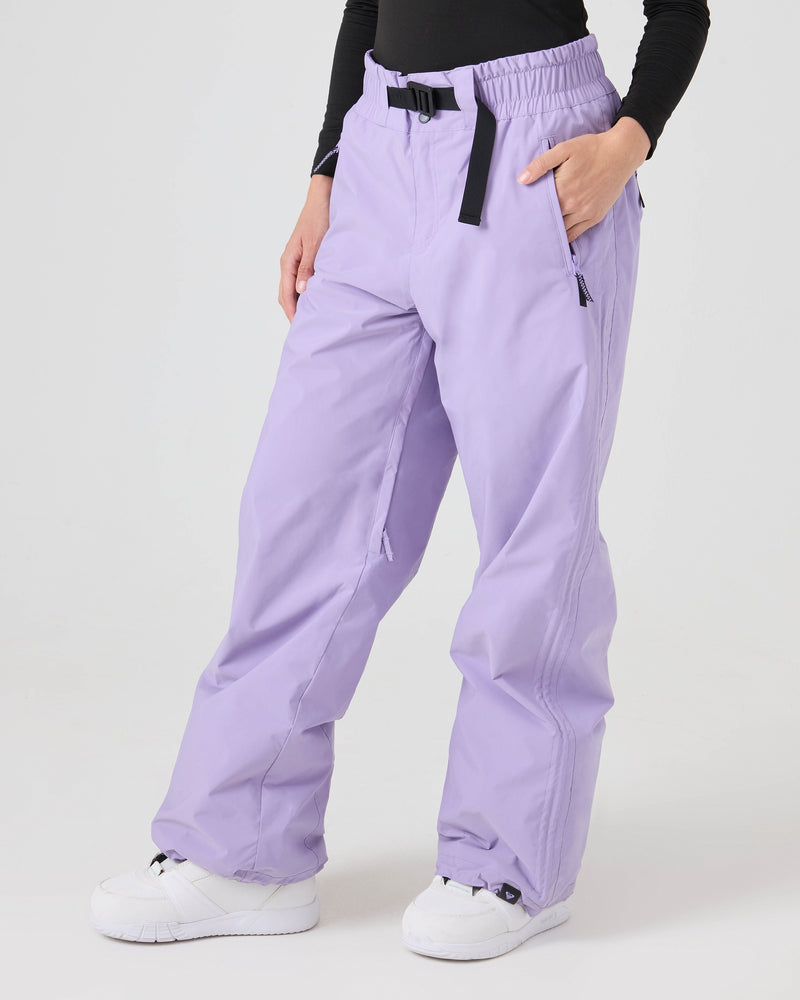 A person wearing Roxy Bleeker Pants in Sunlit Allium—a light purple women’s snow pant featuring 10K/5K protection and venting—paired with a black long-sleeve top and white sneakers, stands with one hand in their pocket against a plain white background.