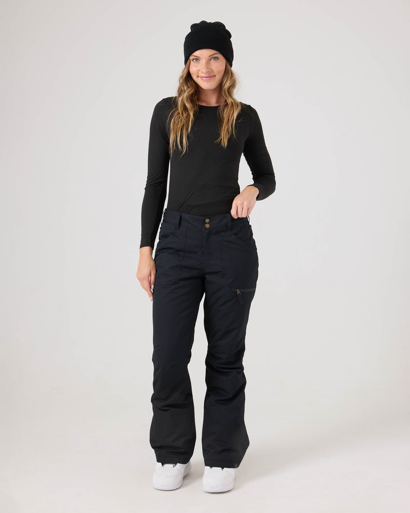 A smiling woman with long blonde hair stands against a solid white background, wearing a black beanie, a black long-sleeve shirt, and the Roxy Nadia Pants—black women’s snow pants crafted from durable corduroy fabric. Her hand rests on the pocket of her waterproof Nadia Pants. She completes her look with white sneakers.