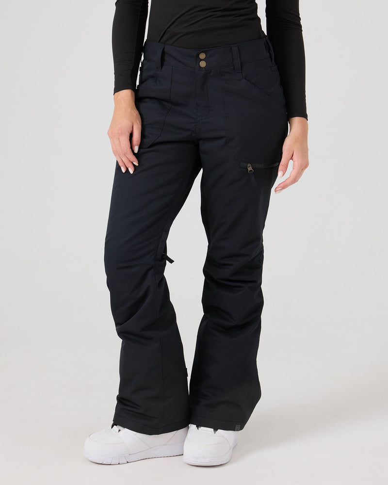 A person is wearing the Roxy Nadia Pants—black corduroy women’s snow pants featuring side and cargo pockets—along with a long-sleeve black shirt and white sneakers, standing against a plain white background.