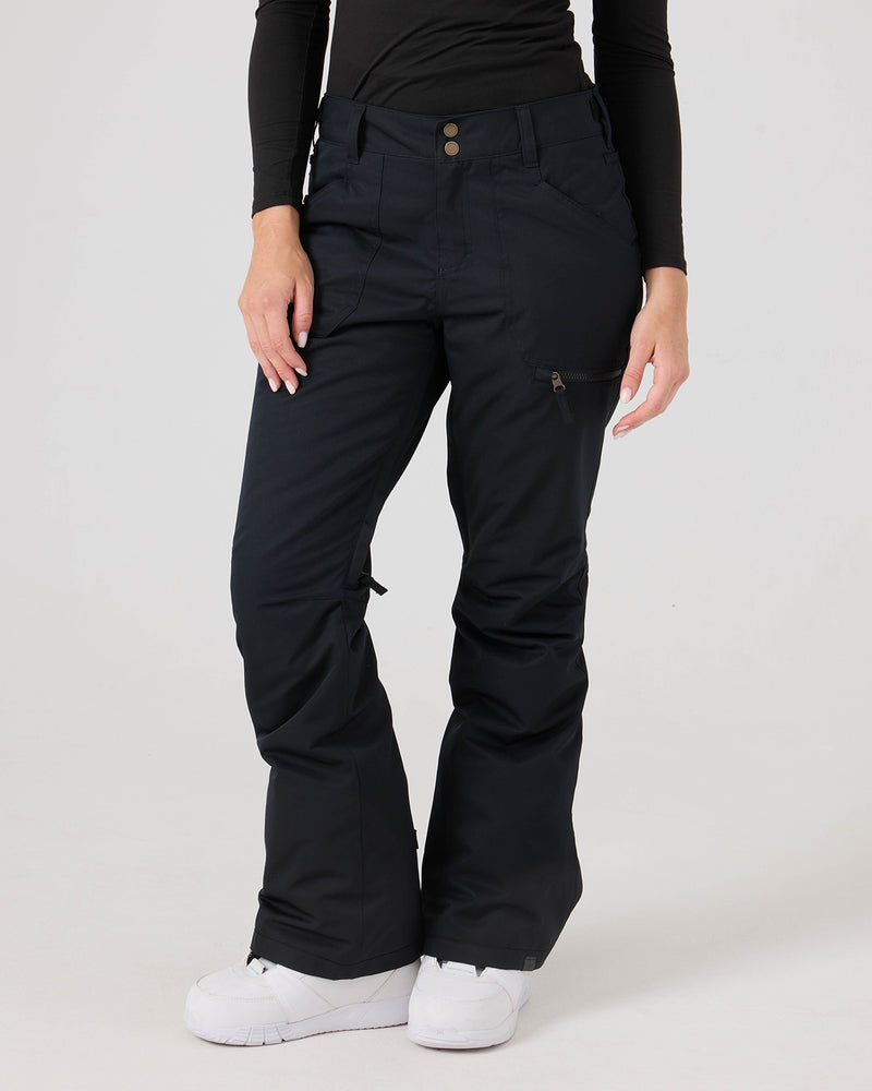 A person is seen standing against a plain background wearing Roxy’s Nadia Pants in black—women’s snow pants crafted from waterproof corduroy fabric, featuring pockets and zippers—with a long-sleeve black shirt and white sneakers. The upper part of the face is not visible.