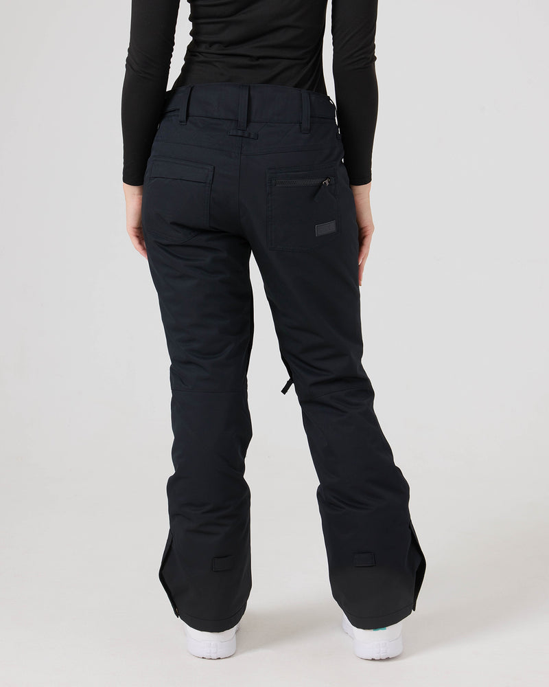 A person wearing Roxy’s Nadia Pants—black women’s snow pants with back pockets and flared legs that cover most of their white shoes—and a black long-sleeve top, stands with their back to the camera.
