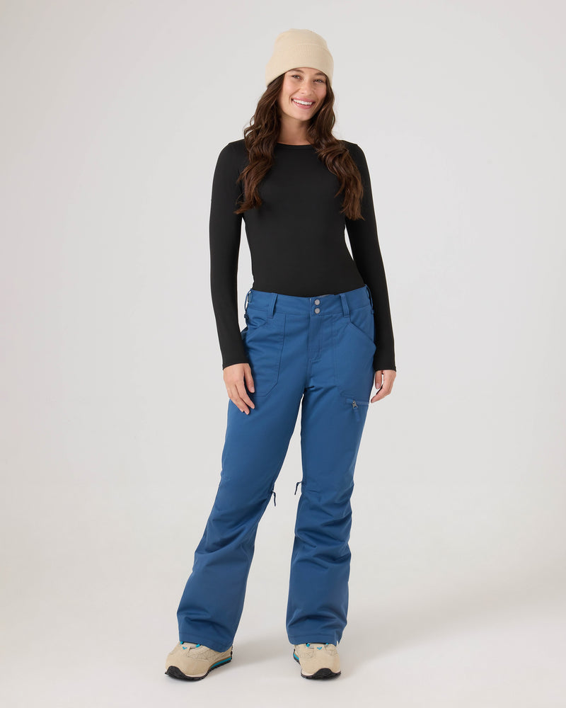 A woman stands smiling with her hands at her sides against a plain light background, wearing a beige beanie, a black long-sleeve top, and blue Roxy Nadia Pants—women’s snow pants crafted from recycled polyester.