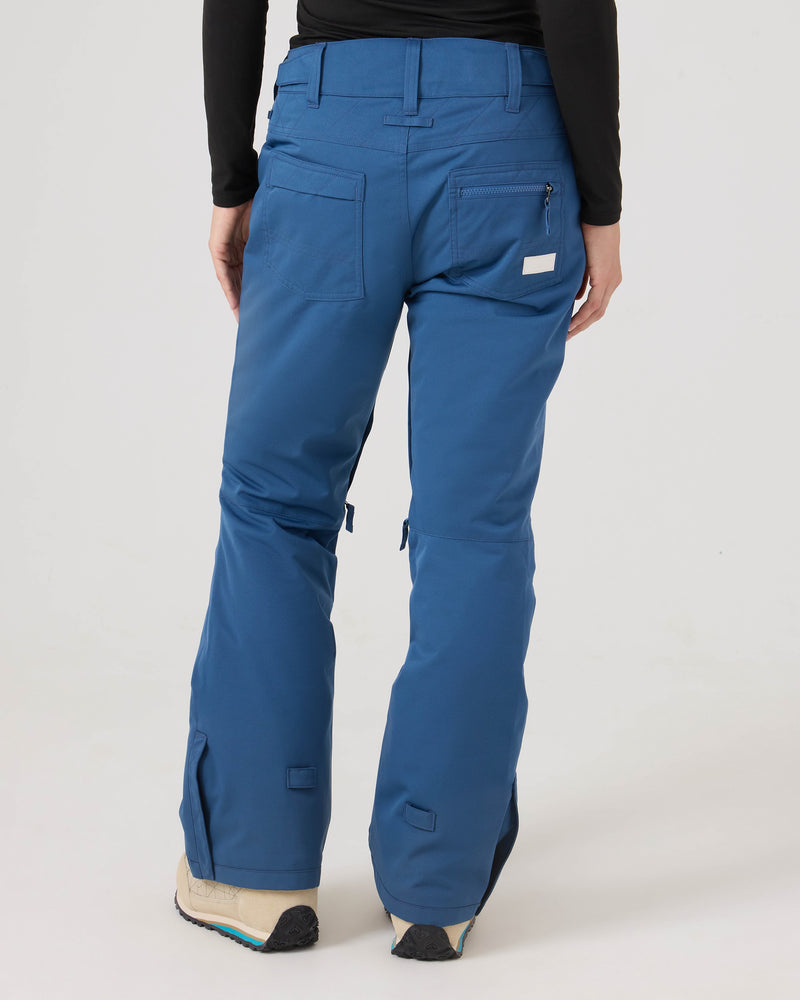 A person is shown from the back wearing blue Roxy Nadia Pants and beige shoes. These women’s snow pants, made from recycled polyester, feature back pockets, zippered details, and flared legs. The background is plain and light-colored.