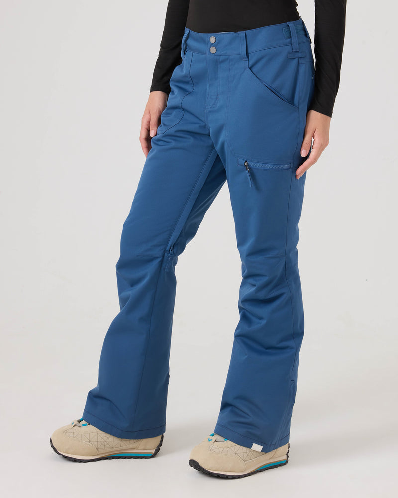 A person wearing Roxy Nadia Pants in Ensign Blue, crafted from recycled polyester with a waterproof-breathable design for comfort and protection, styled with a black long-sleeve shirt and beige shoes featuring blue accents, stands against a plain light background.