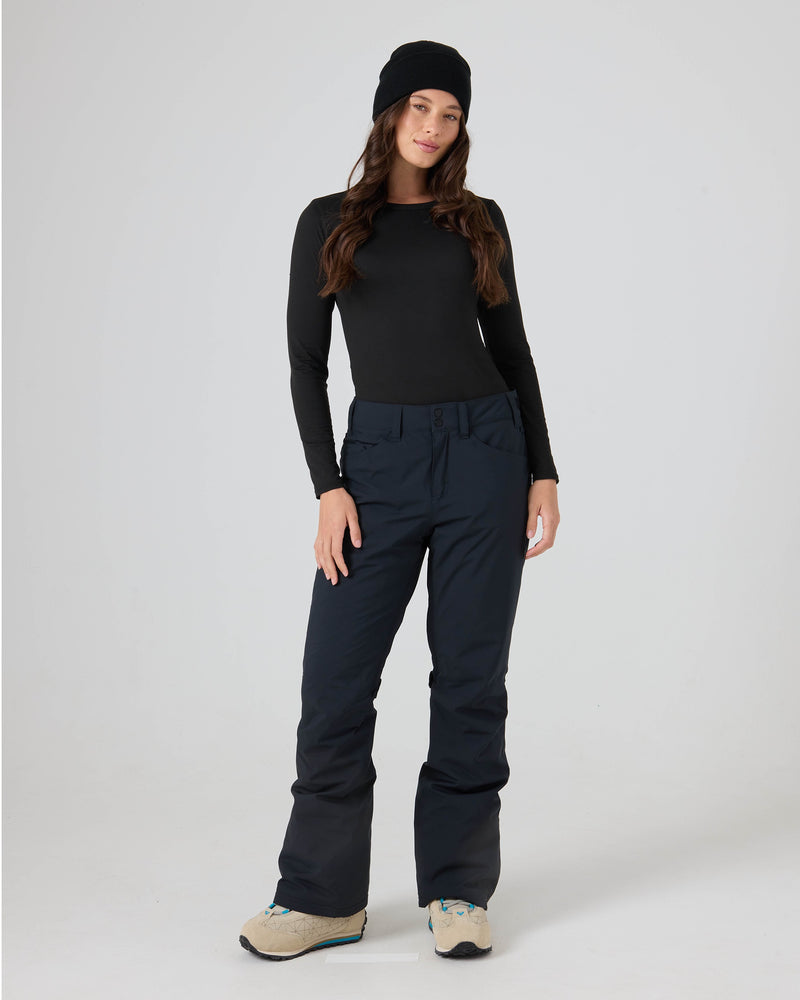A person stands against a plain light background, facing forward with a relaxed expression, dressed in a black beanie, black long-sleeve shirt, and the Roxy Backyard Pants—navy women’s snowboard pants crafted from recycled polyester for dry comfort. The outfit is finished with beige sneakers.