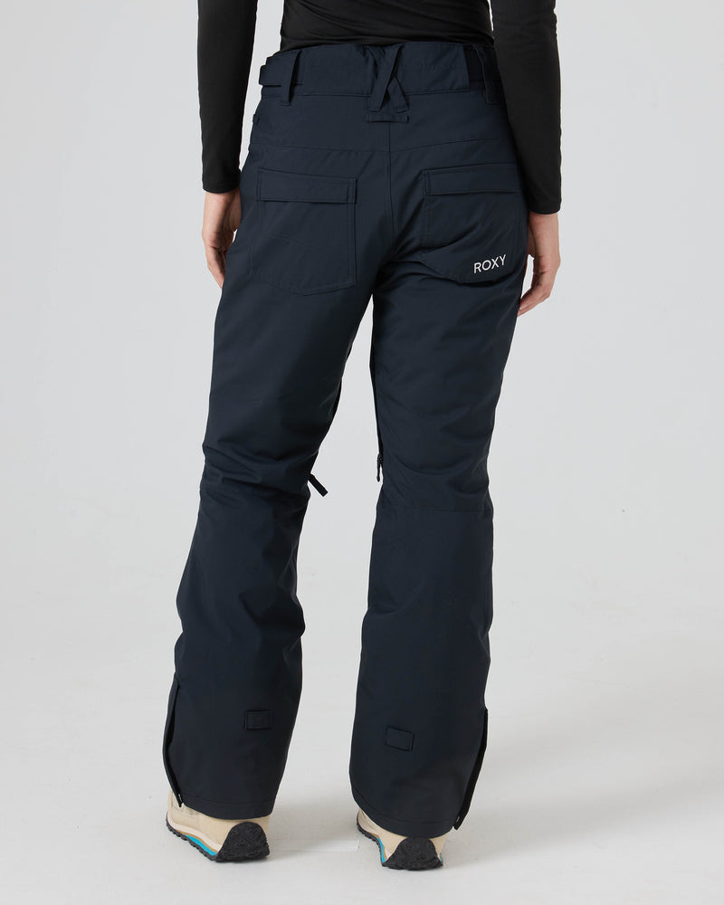 A person is shown from the back wearing dark blue Roxy Backyard Pants for women, crafted from recycled polyester, paired with tan boots on a plain background. The pants feature pockets and visible seams for extra style and dry comfort, while the individual wears a black long-sleeved shirt.