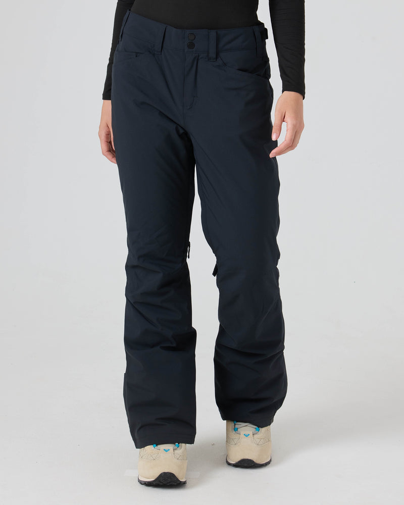 A person is shown from the waist down wearing Roxy Backyard Pants—women’s snowboard pants in black, made for dry comfort from recycled polyester—paired with beige sneakers, standing against a plain white background.