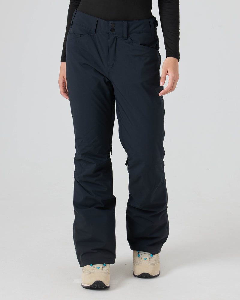 A person wearing Roxy Backyard Pants—black women’s snowboard pants crafted from recycled polyester—paired with a black long-sleeve shirt and white sneakers with blue accents, stands against a plain white background, ready for dry comfort in any winter setting.