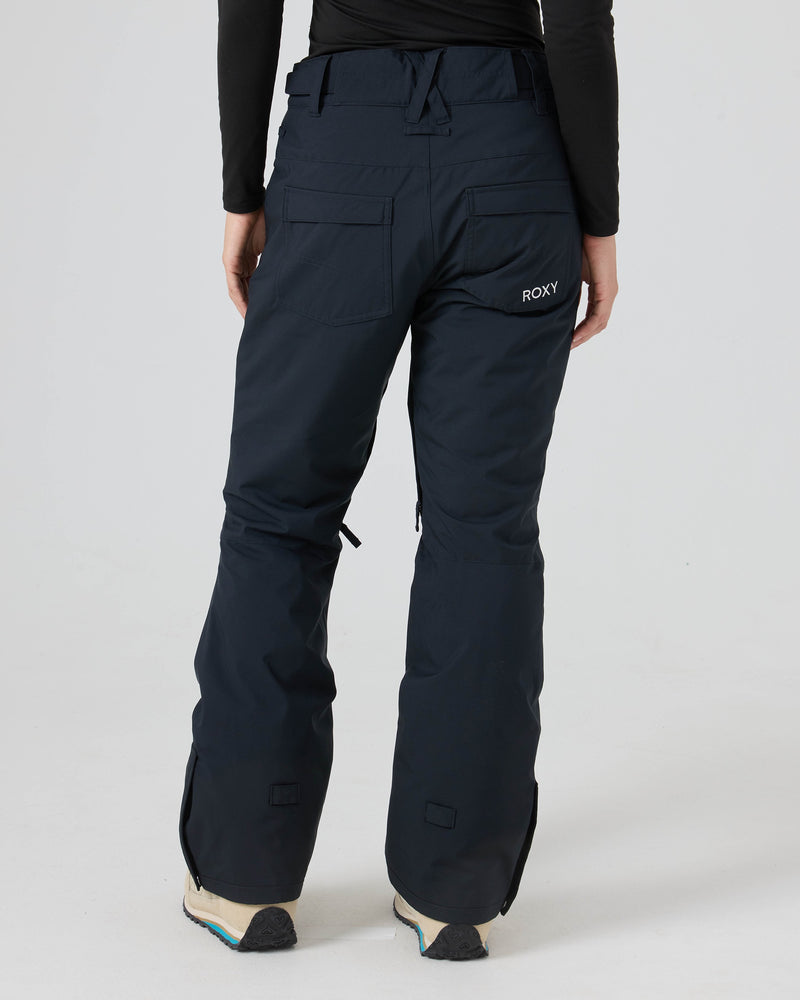 A person stands against a plain white background, shown from the back with the Roxy logo visible on the back pocket, wearing navy blue Roxy Backyard Pants for women. These snowboard pants are made from recycled polyester to provide dry comfort.