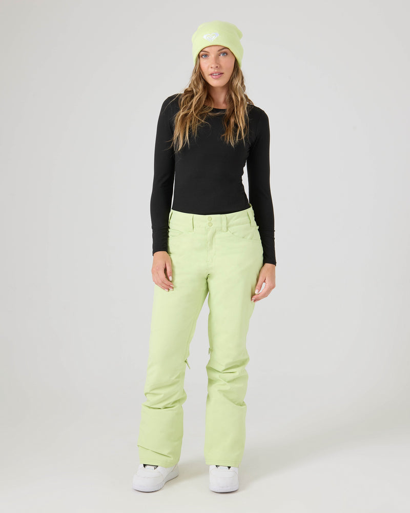 A woman wearing Roxy’s Backyard Pants—warm women’s snowboard pants made from recycled polyester—along with a black long-sleeve shirt and a light green beanie, stands against a plain light background.