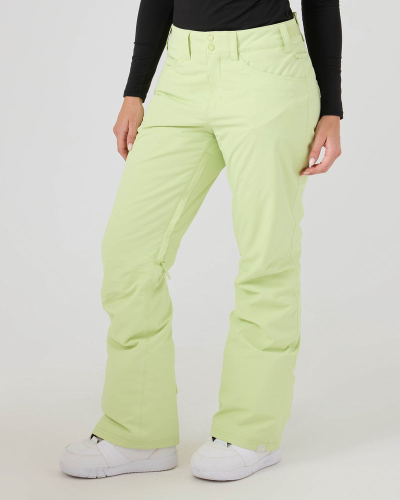 A person is shown from the waist down, standing against a white background, wearing Roxy Backyard Pants in light green—women’s snowboard pants made from recycled polyester—paired with a black long-sleeve top and white sneakers.