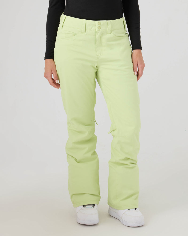 A person wearing pale green Roxy Backyard Pants and a black long-sleeve top stands against a plain white background, with only their body from the shoulders down visible. The loose-fitting women’s snowboard pants are made from recycled polyester for warmth, and the individual completes the look with white sneakers.