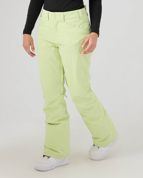 A person is wearing Roxy Backyard Pants in Lily Green—women’s snowboard pants made from warm, recycled polyester—paired with a long-sleeve black top and white shoes, standing against a plain light background. Their face is not visible.