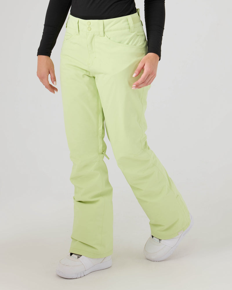 A person is wearing Roxy Backyard Pants in Lily Green—women’s snowboard pants made from warm, recycled polyester—paired with a long-sleeve black top and white shoes, standing against a plain light background. Their face is not visible.