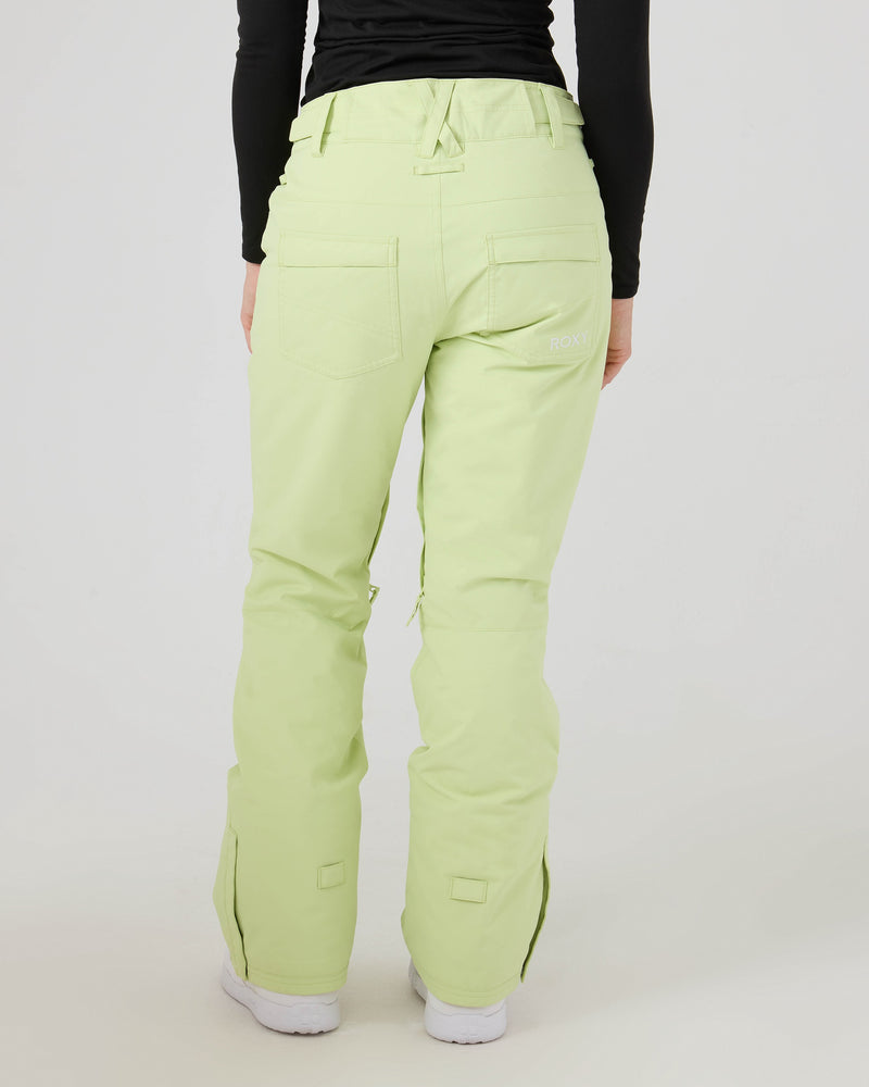A person is seen from behind wearing ROXY Backyard Pants in light green—warm snowboard pants made from recycled polyester, designed with back pockets, prominent seams, and the ROXY logo on the right. The outfit is completed with a black long-sleeve top and white shoes.