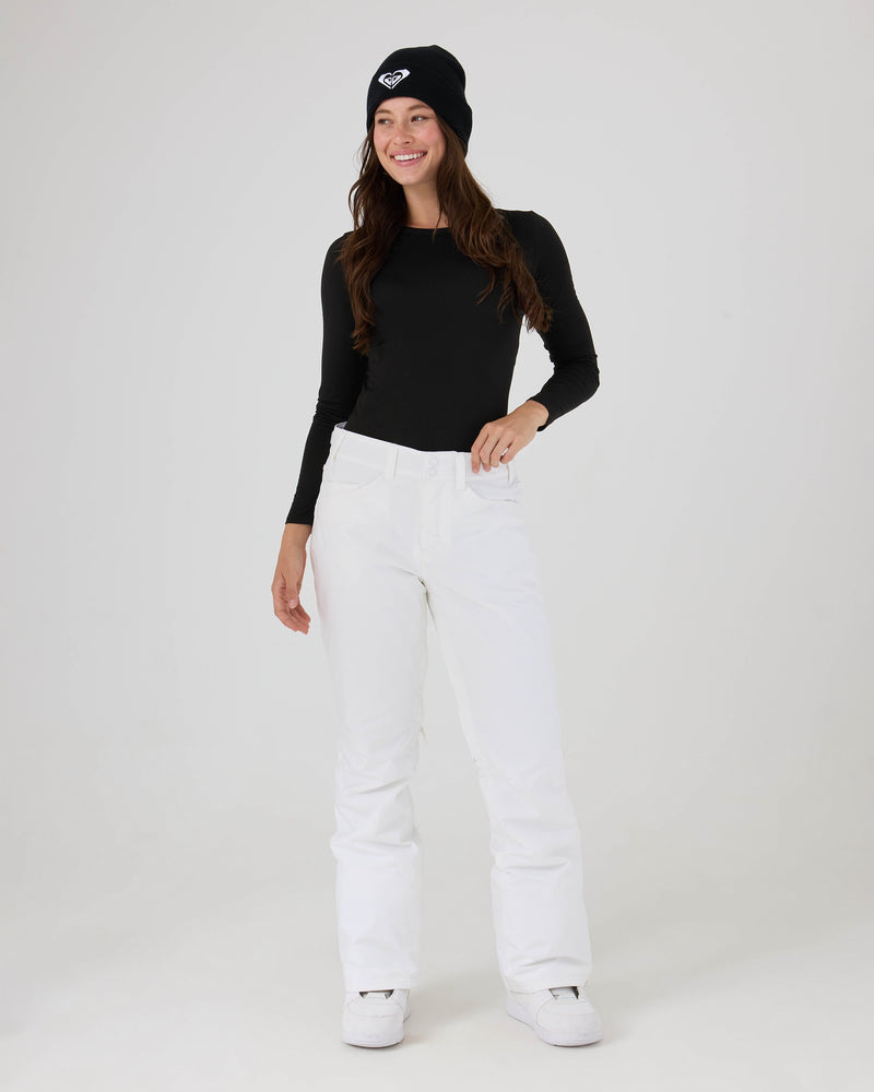 A woman in a black long-sleeve top, white Roxy Backyard Pants made from recycled polyester with 10K/5K protection, white shoes, and a black beanie stands smiling against a plain white background.