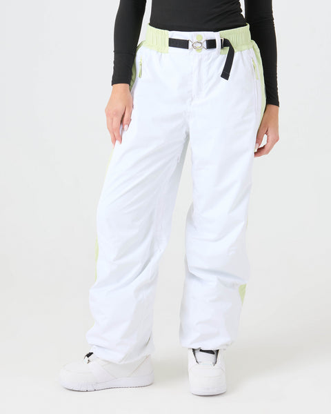 A person is wearing Roxy Chloe Kim Beta Pants in Bright White, accented with light green and offering 15K protection, styled with a black belt, a black long-sleeve top, and white shoes, standing against a plain light background.
