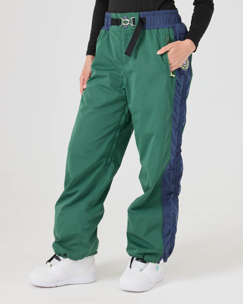 A person is wearing Roxy Chloe Kim Beta Pants in Dkgreen, styled with a black belt, a black long-sleeve top, and white sneakers, standing against a plain light background.