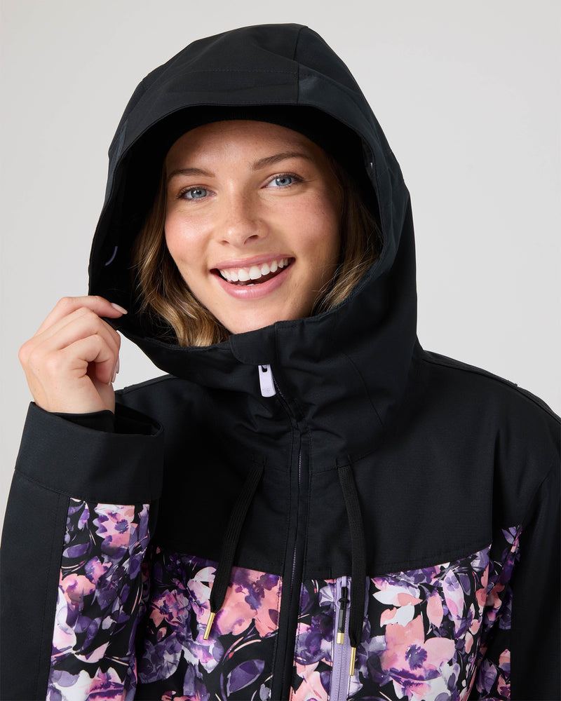 A smiling person models the Roxy Presence Parka Jacket—a women’s snow jacket distinguished by its sleek black hood and eye-catching floral pattern on the chest and sleeves—holding the edge of the hood with one hand, poised to embrace winter with both style and comfort.
