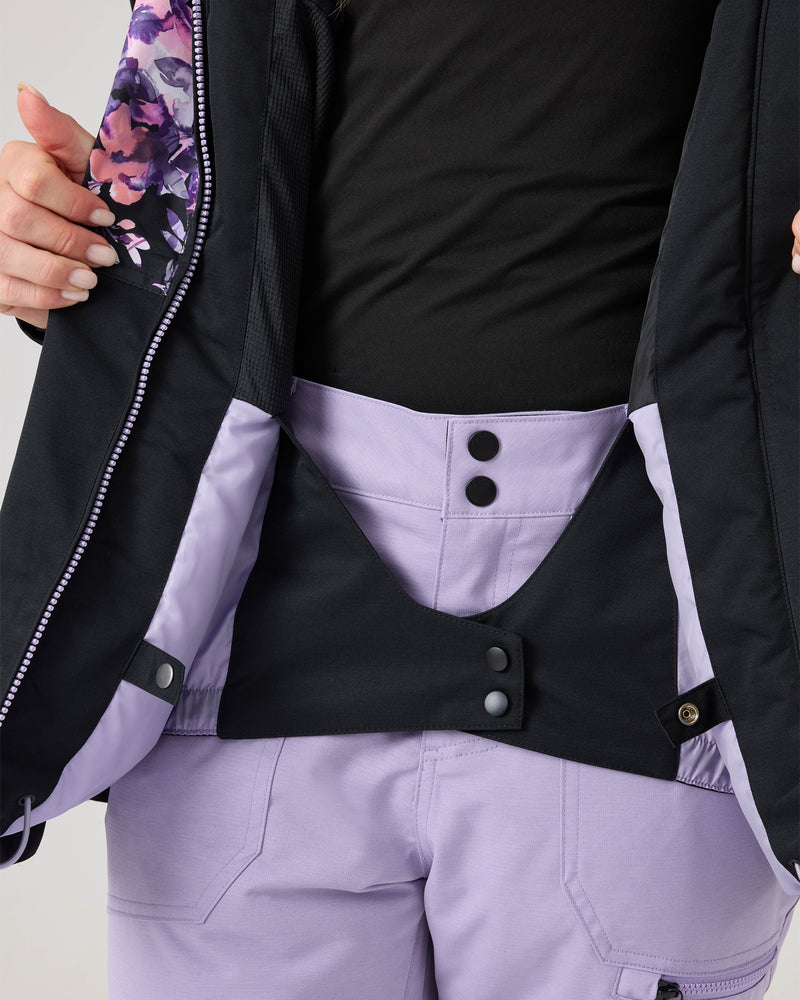 A person wearing a black top and lavender snow pants opens a Roxy Presence Parka Jacket in black with a purple floral lining to reveal the inner waistband and button details of the pants, emphasizing the Presence Parka Jacket’s impressive 15K/15K performance that delivers superior protection on the slopes.
