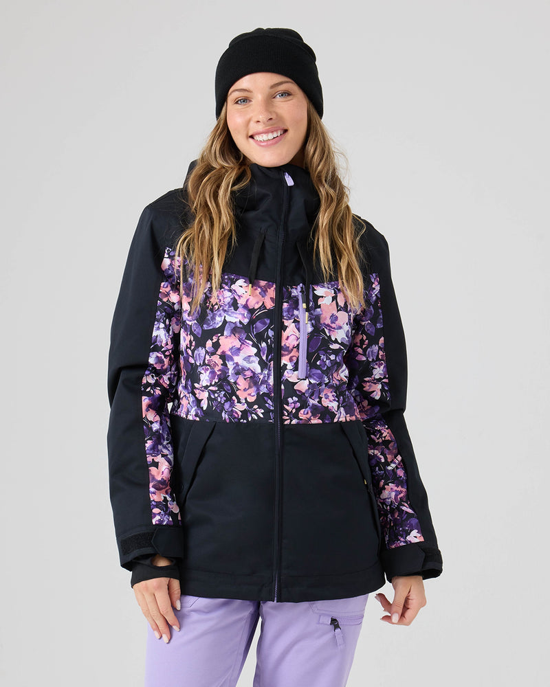 A smiling woman wearing a black beanie, the Roxy Presence Parka Jacket in black, and light purple pants stands against a plain light gray background.