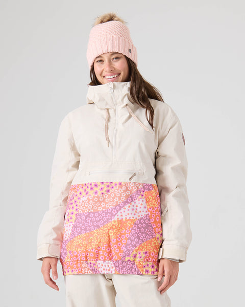 A person wearing the Roxy Downtown Anorak in Petal Power—a cream print-blocked pullover jacket with a vibrant pink and orange floral pattern on the front—and a pink knit beanie, smiling against a plain light background.